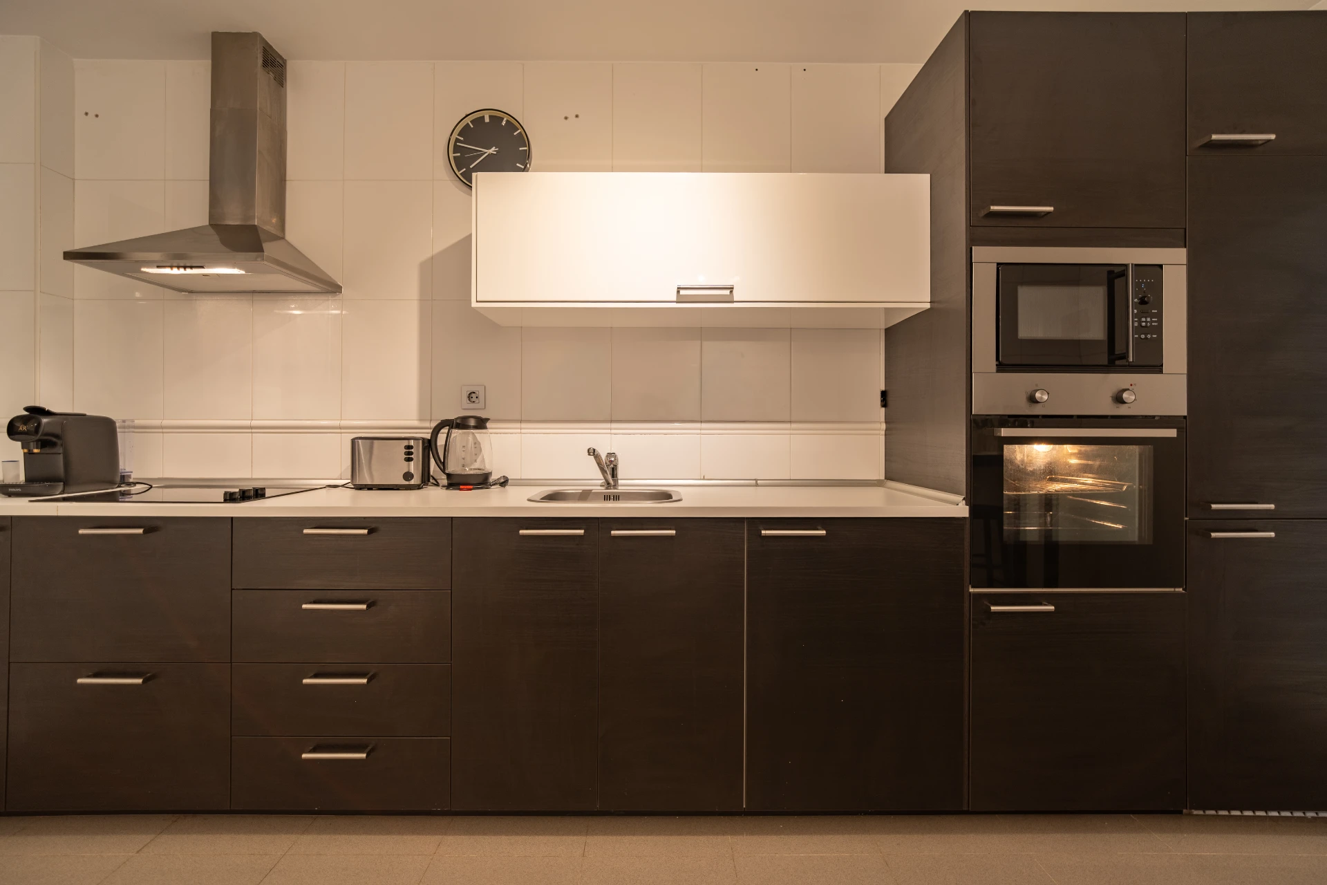 Modern kitchen with dark cabinetry, stainless steel appliances, and a white overhead cabinet, featuring a coffee maker, kettle, and clock, reflecting comfort and convenience for vacation rental guests in Marbella.