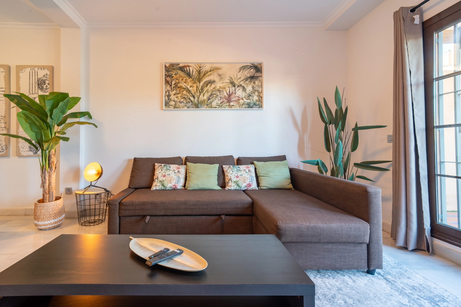 Cozy living area in a Marbella vacation rental featuring a brown sectional sofa with colorful cushions, a decorative plant, a modern coffee table with a remote, and a wall art piece depicting tropical foliage.
