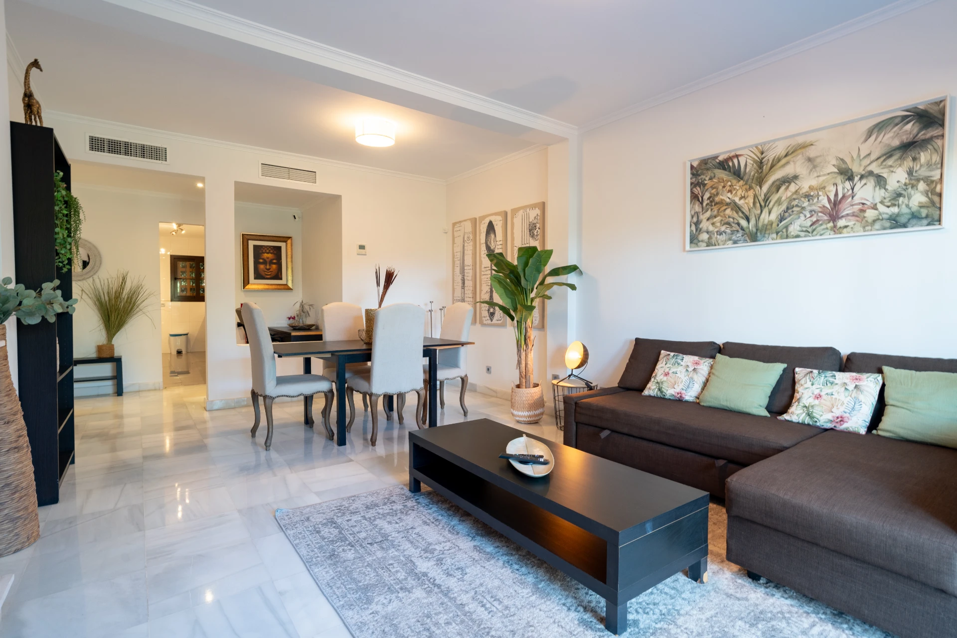Modern living room in Marbella apartment, featuring a cozy brown sofa with decorative pillows, a black coffee table, elegant dining area, tropical artwork, and lush indoor plants.