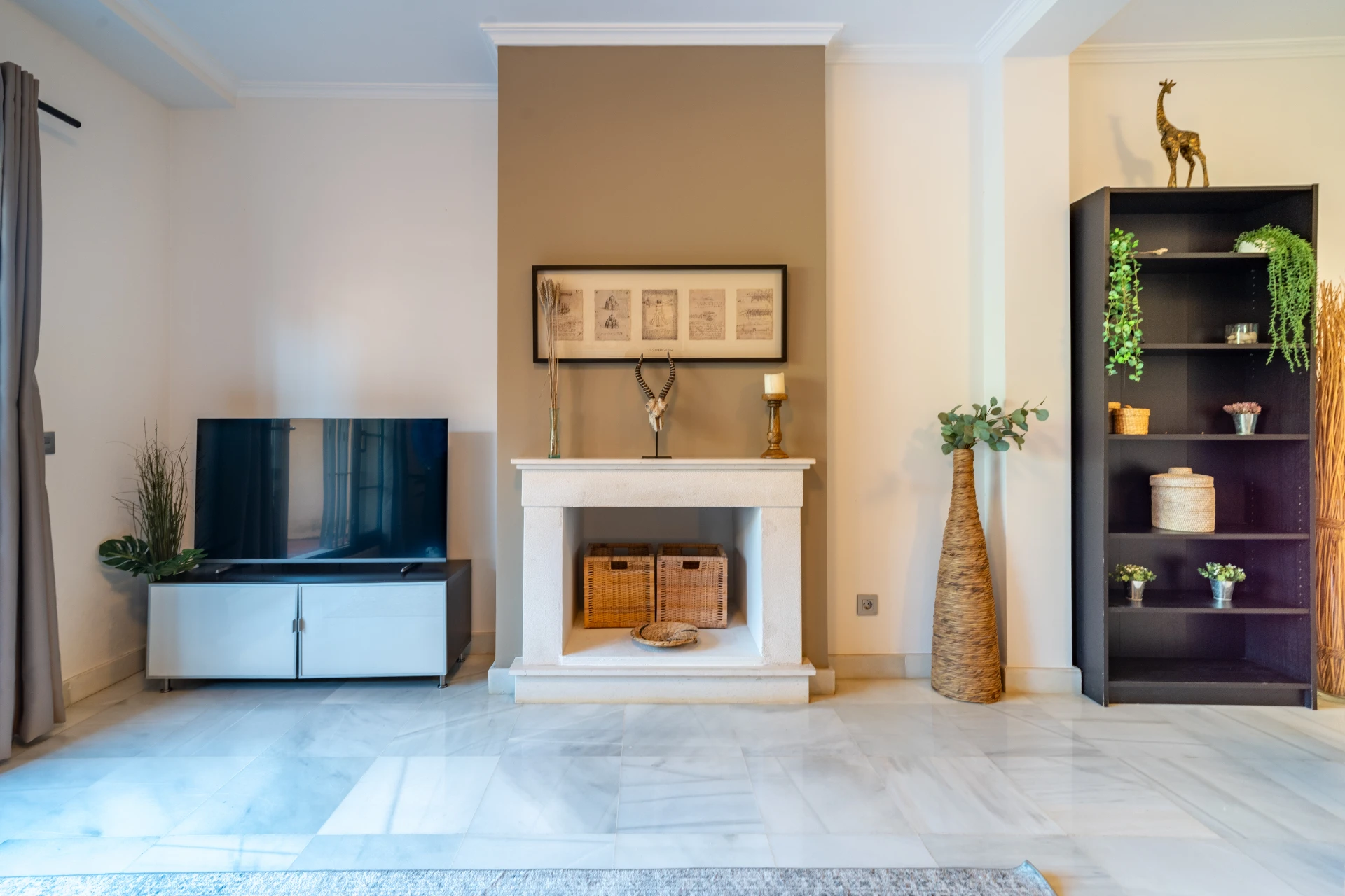 Modern living room featuring a television, decorative fireplace with art and candles, stylish shelving unit with plants and baskets, and elegant marble flooring, ideal for a comfortable stay in Marbella.