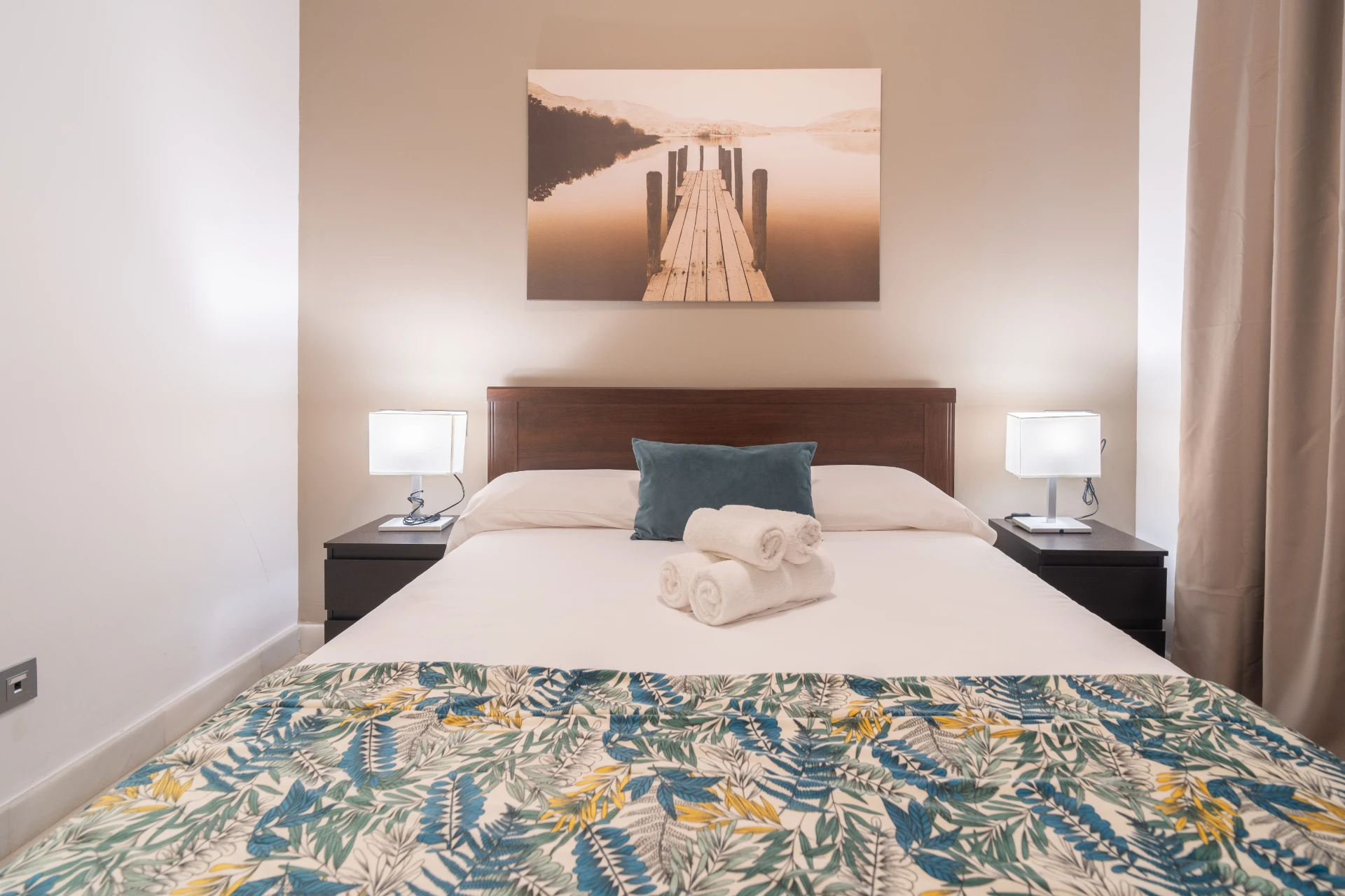 Cozy bedroom with double bed, decorative bedspread featuring tropical leaves, bedside lamps, and a wall art of a serene dock over water, ideal for a relaxing stay in Marbella.