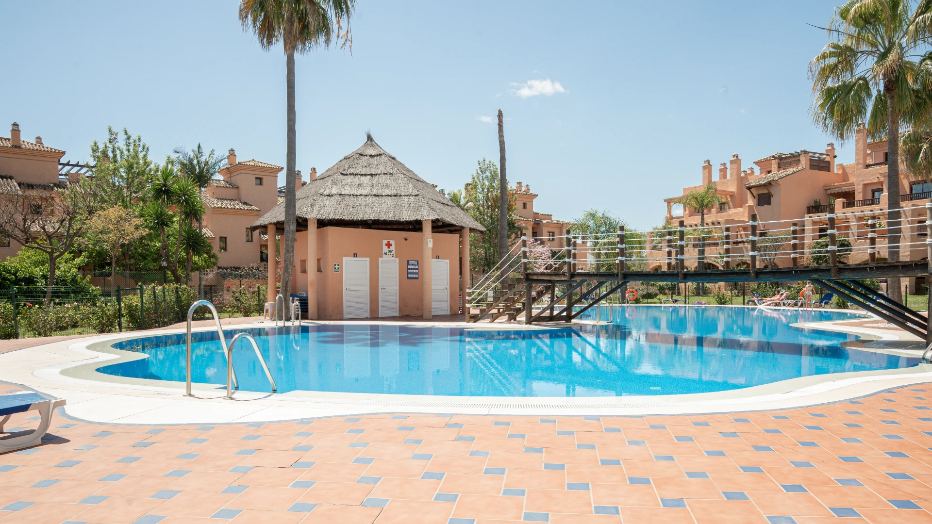 Community swimming pool with sun loungers, surrounded by gardens and palm trees, featuring a thatched-roof gazebo and modern apartment buildings in Marbella, Spain.